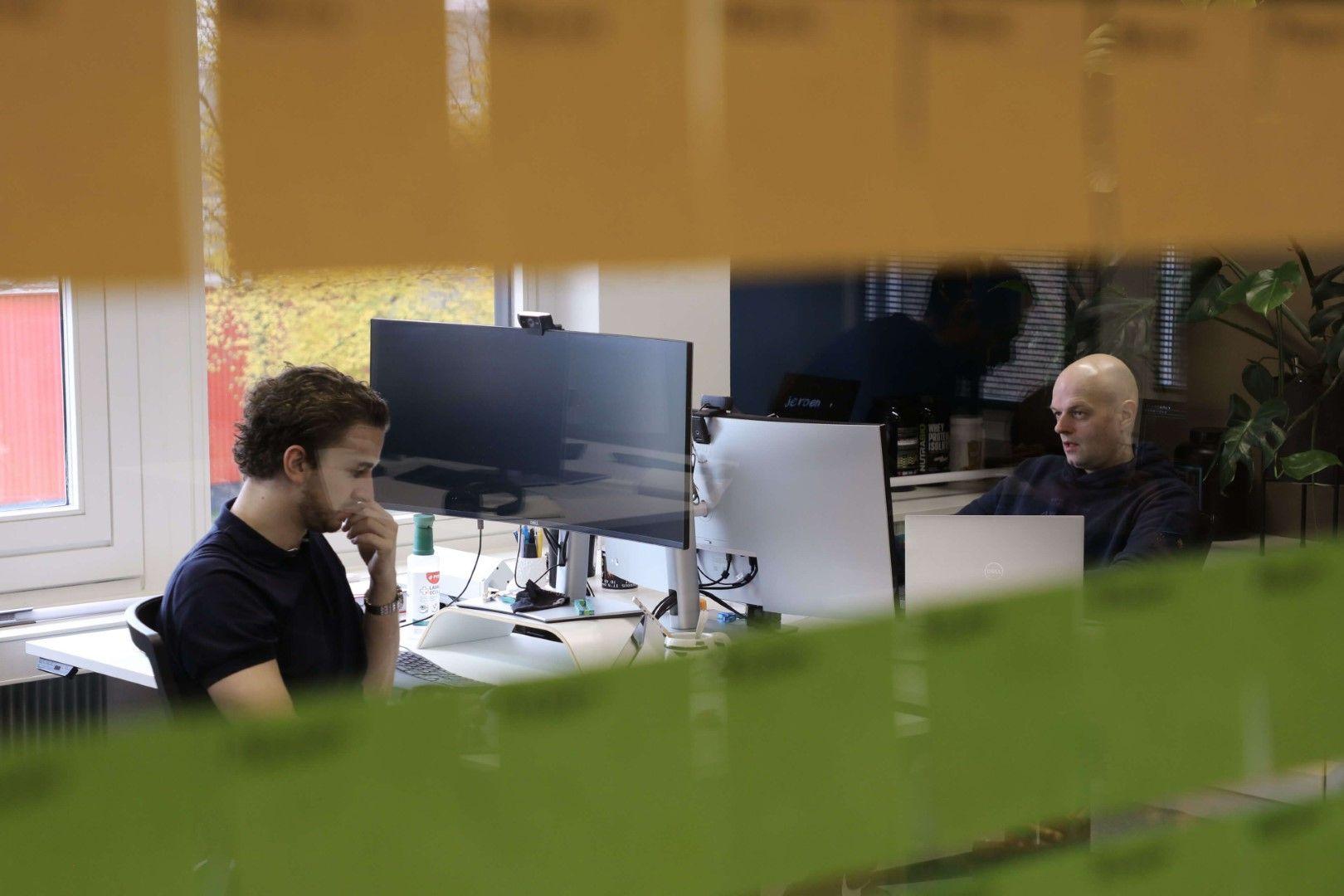 Tim en Jeroen aan het werk achter hun bureaus op kantoor, gefotografeerd door gekleurd glas