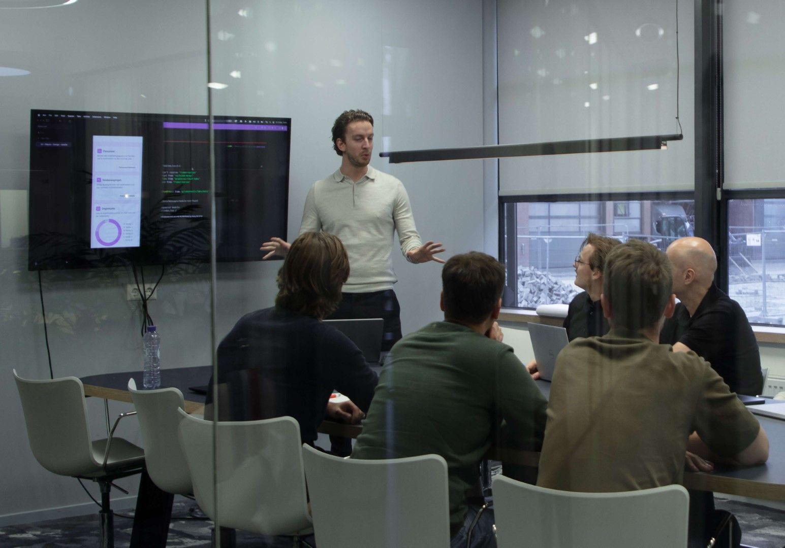 Tim presenteert aan collega's in een vergaderruimte, met code en een app op het scherm