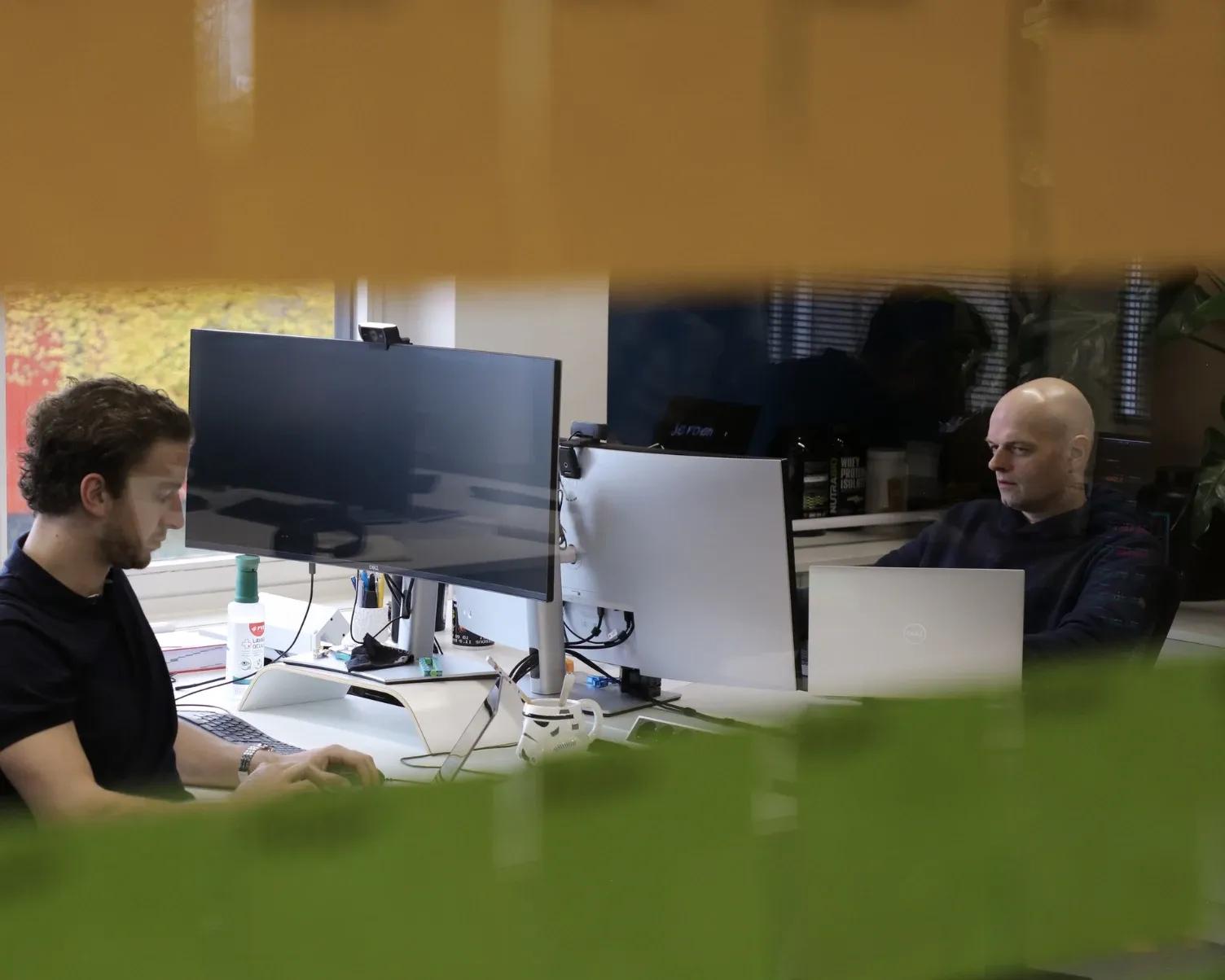 Tim en Jeroen aan het werk achter hun bureaus op kantoor, gefotografeerd door gekleurd glas